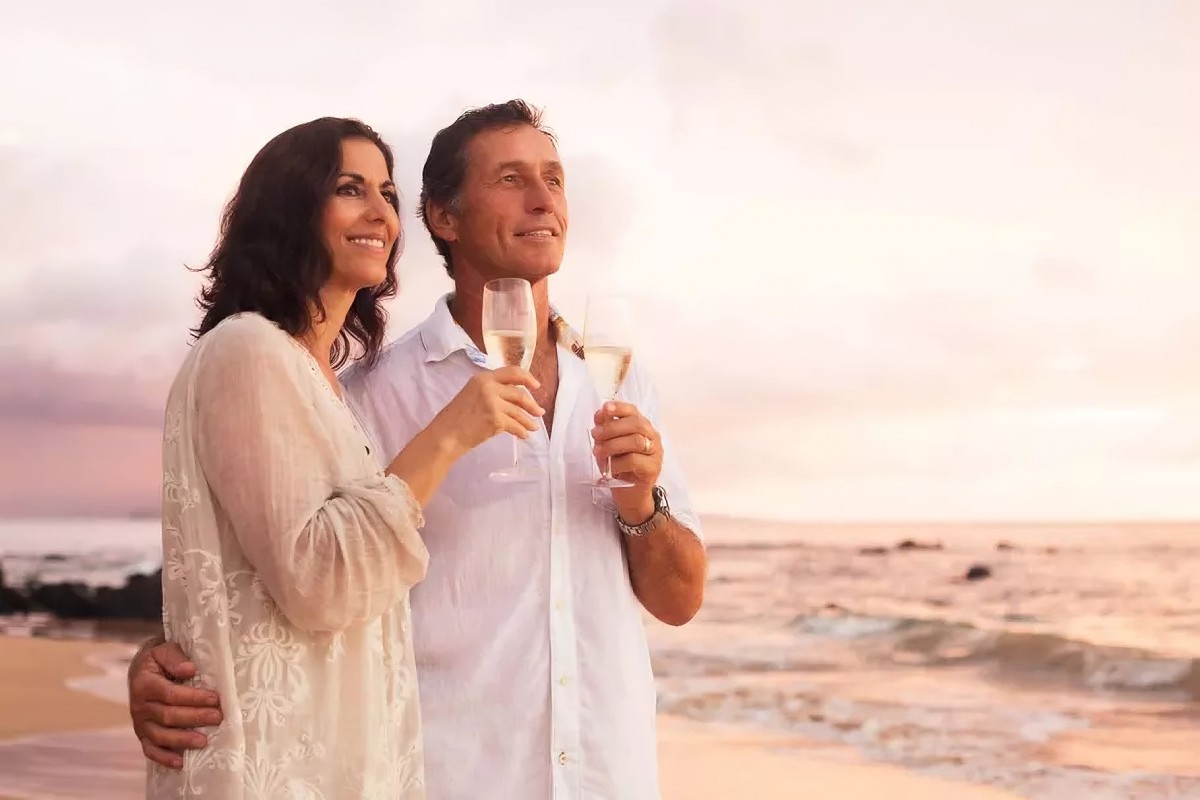 Un couple mature trinque au champagne sur une plage au coucher du soleil pour célébrer leurs 14 ans de mariage, symbolisant les noces de plomb et la solidité de leur union.
